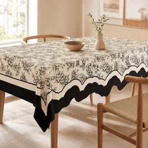 May include: A rectangular tablecloth featuring a cream-colored base with a black floral pattern. The edges are finished with a scalloped black border. A beige ceramic bowl and a small vase with green sprigs sit on the surface of the table.