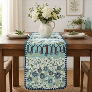 May include: A rectangular table runner featuring a light blue background with a pattern of white and dark blue flowers, brown tree trunks, and green foliage. The runner has a scalloped dark blue border and is draped over a wooden table.