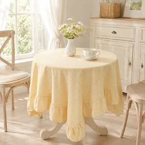 May include: A round table covered with a yellow and white gingham tablecloth featuring a ruffled edge. On the table sits a white ceramic vase filled with white daisies and a white ceramic teacup on a matching saucer, all set on a pedestal base.