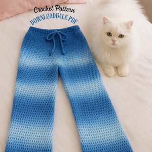 Pode incluir: Calças de crochê com gradiente azul e cintura com cordão. O texto "Crochet Pattern DOWNLOADABLE PDF" é visível. Um gato branco e fofo está ao lado das calças em uma superfície branca.