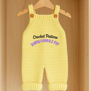 Op de afbeelding: Een gele gehaakte baby-overall met bruine knopen op de bandjes. De tekst "Crochet Pattern DOWNLOADBALE PDF" is zichtbaar op de voorkant. De overall hangt aan een houten kleerhanger in een houten kast.