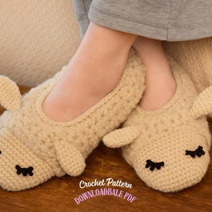 May include: A pair of beige crochet bunny slippers with black embroidered eyelashes and small ears. The slippers are on a wooden surface. The text "Crochet Pattern" and "DOWNLOADBALE PDF" are visible.