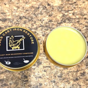 Op de afbeelding: Twee ronde, goudomrande blikken met gietijzeren kruidencompound. Eén blik is open en onthult een lichtgele balsem. Het gesloten blik heeft een zwart etiket met de tekst "JOES CAST IRON RESTORE" en "CAST IRON SEASONING COMPOUND."