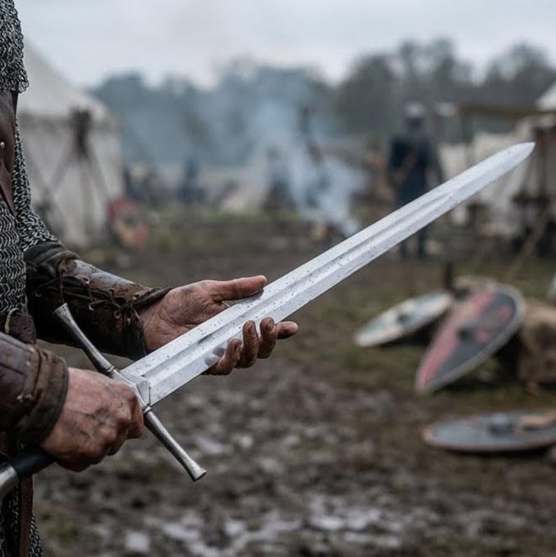 Op de afbeelding: Een lang, zilveren zwaard met een kruisvormige handgreep wordt vastgehouden. Het lemmet is lang en recht, met een punt. De achtergrond toont een middeleeuws kampement met tenten en schilden. Het zwaard is ongeveer 90 cm lang.