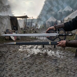 Op de afbeelding: Een lang, zilveren zwaard met een zwart handvat en een decoratief schild met gouden accenten. Het zwaard wordt boven een zwarte schede met gouden strepen gehouden. De sc&egrave;ne speelt zich buiten af met tenten op de achtergrond.