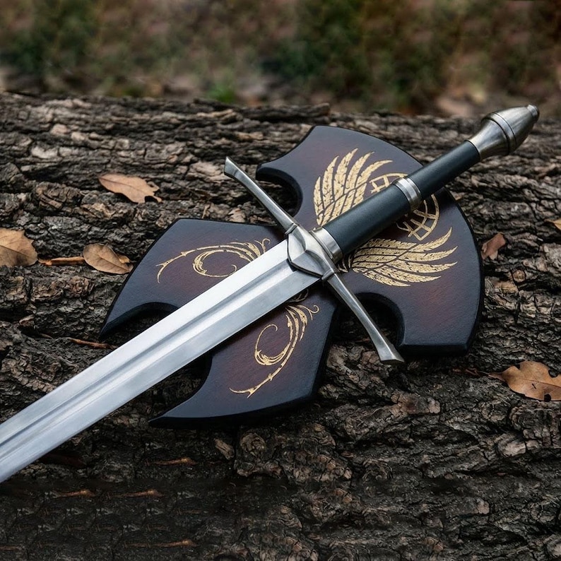 Op de afbeelding: Een lang, zilveren zwaard met een zwarte handgreep en kruisbescherming rust op een houten schild. Het schild is donkerbruin met goudkleurige vleugel- en krulontwerpen. Het zwaard heeft een hoogglans gepolijst lemmet.