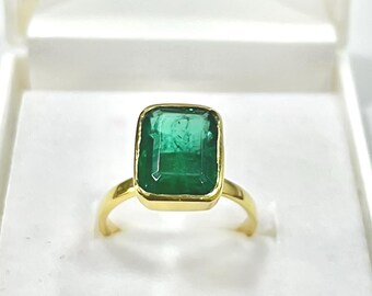 Op de afbeelding: Een gouden ring met een grote, rechthoekige smaragd. De smaragd heeft een levendige groene kleur en is gezet in een gouden band. De ring wordt gepresenteerd in een witte sieradendoos.