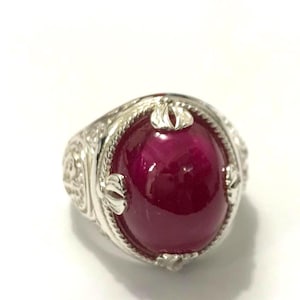 Può includere: Un anello d'argento con una gemma ovale di colore rosso intenso. L'anello presenta dettagli decorativi attorno alla gemma e sulla fascia. La gemma è il punto focale, incastonata in una montatura d'argento decorativa. L'anello è su una superficie bianca.