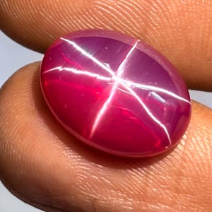 May include: Oval-shaped, polished ruby gemstone with a six-rayed star effect. The gem is a deep pinkish-red colour, with a bright white star pattern across its surface. The gemstone is held in a hand.