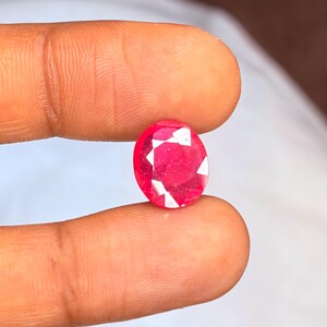 May include: A faceted, oval-shaped ruby gemstone. The ruby is a vibrant red color with white facets that catch the light. The gemstone is held between two fingers, showcasing its size and brilliance.