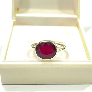 May include: A silver ring with a large, round, deep red gemstone. The ring is set in a simple, polished silver band and is displayed in an open, off-white jewelry box. The gemstone is the focal point.