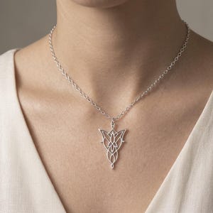 May include: A silver-colored pendant necklace featuring a stylized floral design. The pendant hangs from a delicate chain, creating a simple yet elegant accessory. The necklace is worn against a cream-colored top.