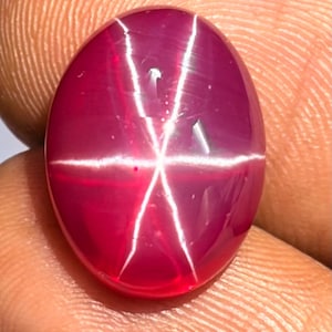 May include: A polished, oval-shaped gemstone in a deep red hue, displaying a six-rayed star pattern. The star effect is due to light interacting with internal inclusions. The gem's surface is smooth, highlighting its rich colour.