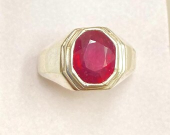 Op de afbeelding: Een zilverkleurige ring met een grote, ovaal geslepen, dieprode edelsteen in een getrapte, geometrische zetting. De ringband is glad en loopt taps toe naar de achterkant, terwijl de centrale zetting het gefacetteerde oppervlak van de steen benadrukt.
