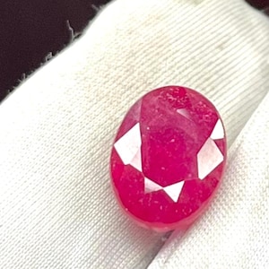 May include: A faceted, oval-shaped ruby gemstone. The ruby is a deep, vibrant red color with white, geometric facets. The gemstone is set against a white, textured background.