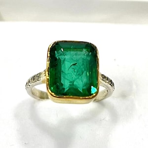 Può includere: Anello con pietra verde rettangolare taglio smeraldo, con cornice dorata e fascia argentata. La pietra verde, di grandi dimensioni, è messa in risalto. La fascia è ornata da piccole pietre scintillanti.