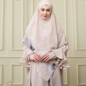 May include: A beige hijab and matching dress. The hijab has a detailed pattern and a contrasting border. The dress has long sleeves with ruffled cuffs. The image includes the text "Ivory Bloom".