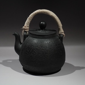 May include: A black cast iron teapot with a textured surface and a woven handle. The teapot has a curved spout and a small lid with a round knob. The handle is made of a light-colored, woven material.