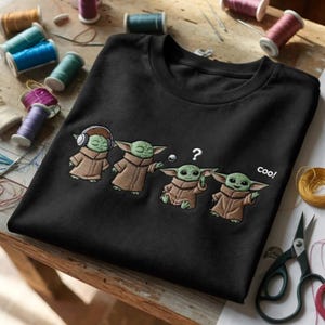 Puede incluir: Camiseta negra con un dise&ntilde;o bordado de cuatro personajes Grogu. Uno lleva auriculares, otro tiene un signo de interrogaci&oacute;n sobre la cabeza y el &uacute;ltimo dice "coo!"