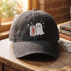 May include: A charcoal gray baseball cap featuring an embroidered design of a white ghost holding a silver soda can with the red text Coke on it. The embroidery includes small white stars and a black bat silhouette above the ghost figure.