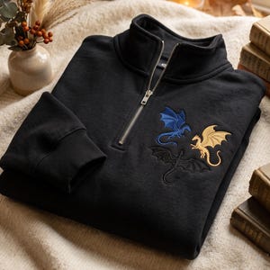 May include: A black quarter-zip sweatshirt featuring an embroidered design of three dragons on the chest. The dragons are rendered in blue, gold, and black thread. The garment has a high collar with a silver-toned metal zipper closure.