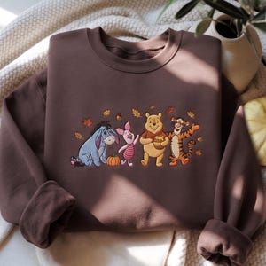 Pode incluir: Uma camisola de gola redonda castanho-escura com um bordado do Ió, Leitão, Winnie the Pooh a segurar um pote de mel e o Tigre. As personagens estão rodeadas no peito por pequenas folhas de outono cor de laranja e amarelas e bolotas.