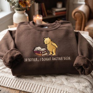 May include: A dark brown crewneck sweatshirt featuring an embroidered graphic of a yellow bear standing next to a stack of two books. Below the graphic, the text reads, "OH BOTHER... I BOUGHT ANOTHER BOOK" in white, capitalized, sans-serif lettering.