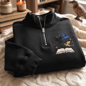 May include: A black quarter-zip sweatshirt featuring an embroidered design on the chest. The embroidery depicts an open book with a blue dragon, a black dragon, and a yellow dragon flying above it, surrounded by small embroidered stars.