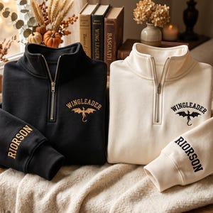 May include: Two quarter-zip sweatshirts, one black and one cream, each featuring an embroidered gold dragon graphic with the text WINGLEADER above it on the chest and the name RIORSON embroidered in a matching color on the sleeve cuff.