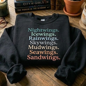 May include: A black sweatshirt with the words "Nightwings, Icewings, Rainwings, Skywings, Mudwings, Seawings, Sandwings" in a gradient of colours. The sweatshirt is on a wooden table with books, a candle, and a small potted plant.