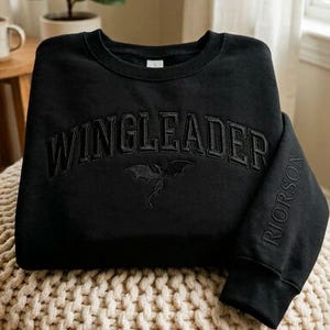 May include: Black crewneck sweatshirt with the word "WINGLEADER" arched across the chest in a raised, textured font. A dragon graphic is below the text. The right sleeve has the name "RIORSON" vertically embroidered. The sweatshirt is folded on a beige pouf.