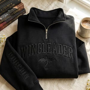 May include: A black quarter-zip sweatshirt featuring the embroidered text WINGLEADER in an arched font across the chest, with a dragon silhouette below. The sleeve features the embroidered text RIORDAN, and the collar has a silver zipper.