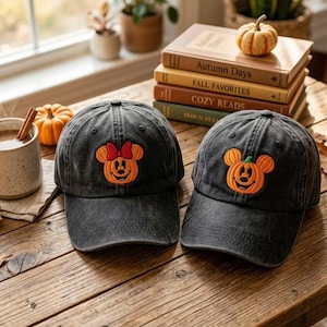 May include: Two charcoal gray baseball caps featuring embroidered pumpkin character designs. One cap displays a pumpkin with a red bow, while the other features a pumpkin with a green stem. Both caps have a curved brim and a structured crown.