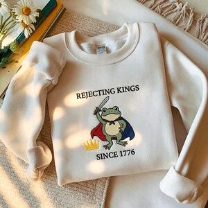 Könnte beinhalten: Ein cremefarbenes Sweatshirt mit Rundhalsausschnitt und einer Grafik eines grünen Frosches, der einen blau-roten Umhang trägt, ein Schwert hält und neben einer gelben Krone steht. Der Text REJECTING KINGS SINCE 1776 ist in Schwarz aufgedruckt.