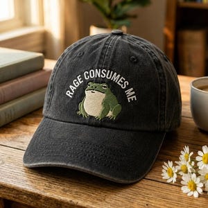May include: A charcoal gray baseball cap featuring an embroidered green frog centered on the front panel. Above the frog, white text is arched in a semi-circle and reads RAGE CONSUMES ME. The cap has a curved brim and a structured crown design.