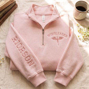 May include: A light pink quarter-zip sweatshirt featuring the embroidered text WINGLEADER above a dragon graphic on the chest and the name RIORSON embroidered vertically down the right sleeve. The garment has a high collar and ribbed cuffs.