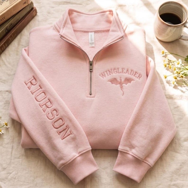 May include: A light pink quarter-zip sweatshirt featuring the embroidered text WINGLEADER above a dragon graphic on the chest and the name RIORSON embroidered vertically down the right sleeve. The garment has a high collar and ribbed cuffs.