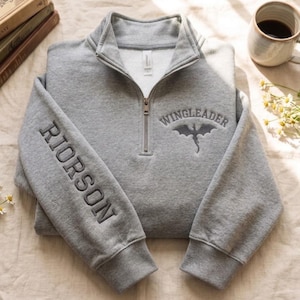 May include: A heather gray quarter-zip sweatshirt featuring the embroidered text RIORSON vertically on the left sleeve and WINGLEADER with a dragon silhouette on the right chest. The garment has a high collar, a metal zipper, and ribbed cuffs.