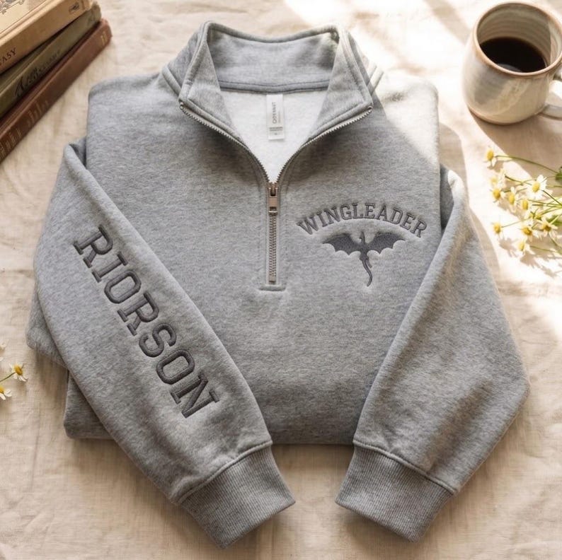 May include: A heather gray quarter-zip sweatshirt featuring the embroidered text RIORSON vertically on the left sleeve and WINGLEADER with a dragon silhouette on the right chest. The garment has a high collar, a metal zipper, and ribbed cuffs.