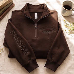 May include: A dark brown quarter-zip sweatshirt featuring the embroidered text RIORSON vertically on the right sleeve and WINGLEADER with a dragon graphic on the chest. The garment has a high collar, a metal zipper, and ribbed cuffs.