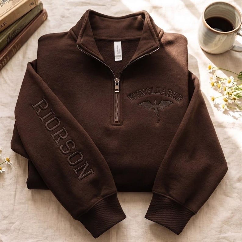 May include: A dark brown quarter-zip sweatshirt featuring the embroidered text RIORSON vertically on the right sleeve and WINGLEADER with a dragon graphic on the chest. The garment has a high collar, a metal zipper, and ribbed cuffs.