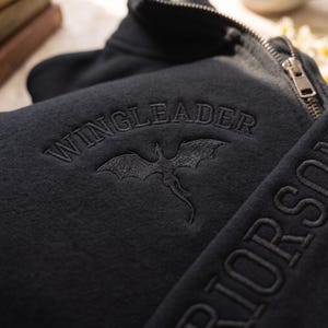 May include: A black zip-up sweatshirt featuring the embroidered text WINGLEADER above a dragon silhouette. The garment includes a metal zipper pull and additional embroidered text visible along the side panel of the fabric.