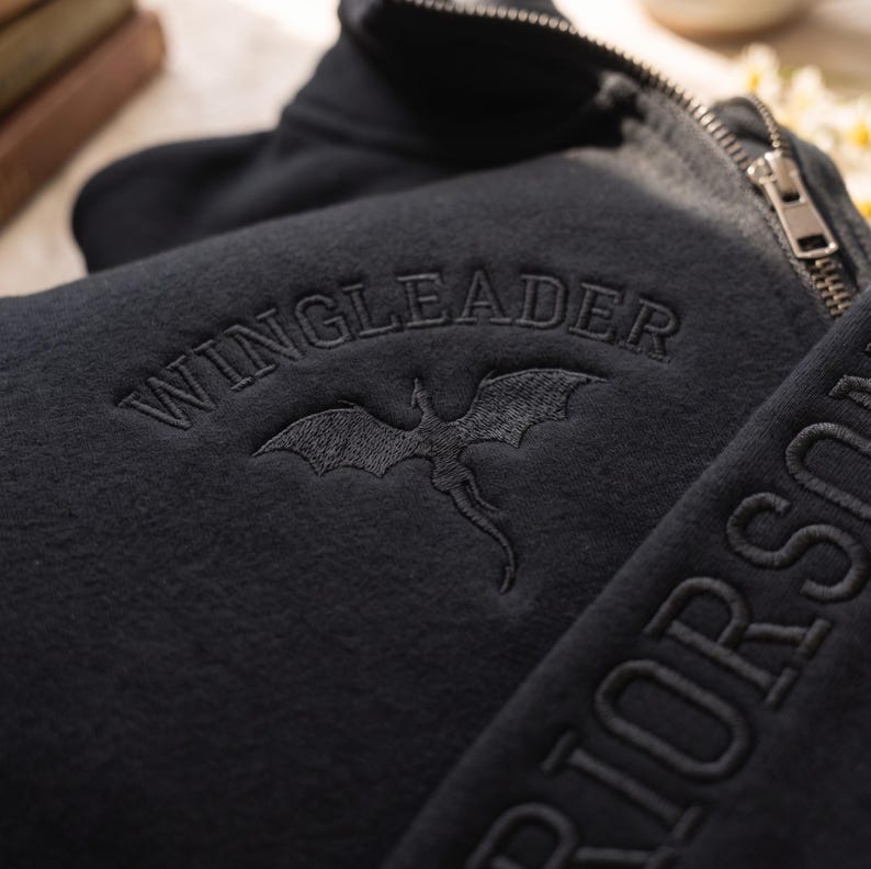 May include: A black zip-up sweatshirt featuring the embroidered text WINGLEADER above a dragon silhouette. The garment includes a metal zipper pull and additional embroidered text visible along the side panel of the fabric.
