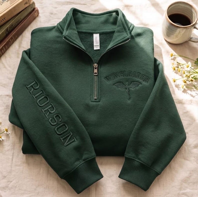 May include: A dark green quarter-zip sweatshirt featuring the embroidered text RIORSON vertically on the right sleeve and WINGLEADER with a dragon graphic on the chest. The garment has a high collar, a metal zipper, and ribbed cuffs.