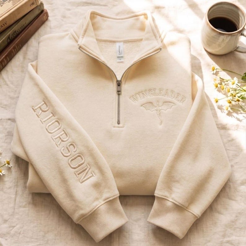 May include: A cream-colored quarter-zip sweatshirt featuring the embroidered text RIORSON on the right sleeve and WINGLEADER with a dragon graphic on the chest. The garment has a metal zipper, ribbed cuffs, and a high collar.