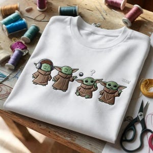 Puede incluir: Camiseta blanca con un dise&ntilde;o bordado de un personaje alien&iacute;gena verde con una t&uacute;nica marr&oacute;n. El dise&ntilde;o incluye la palabra "coo!" y un signo de interrogaci&oacute;n. Suministros de costura rodean la camiseta.