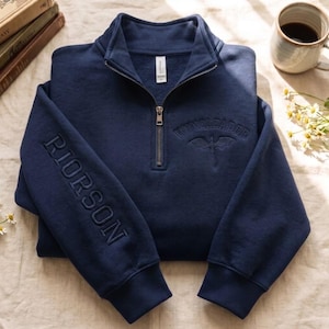 May include: A navy blue quarter-zip sweatshirt featuring the embroidered text RIORSON on the sleeve and WINGLEADER with a dragon graphic on the chest. The garment has a metal zipper, ribbed cuffs, and a high collar, presented in a folded position.