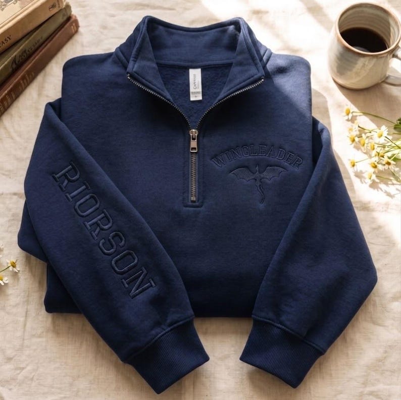 May include: A navy blue quarter-zip sweatshirt featuring the embroidered text RIORSON on the sleeve and WINGLEADER with a dragon graphic on the chest. The garment has a metal zipper, ribbed cuffs, and a high collar, presented in a folded position.