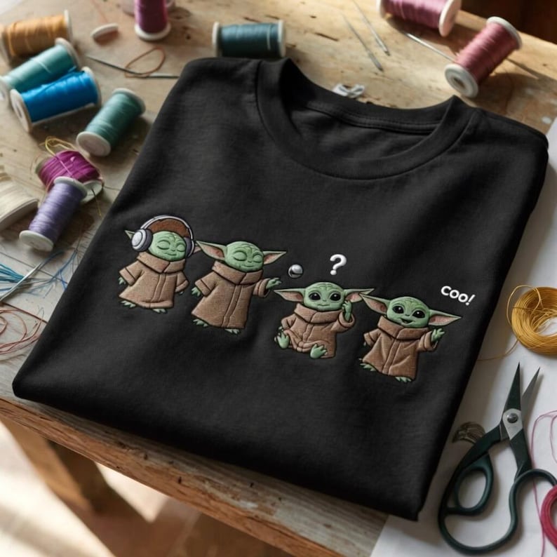 Puede incluir: Camiseta negra con un dise&ntilde;o bordado de cuatro personajes Grogu. Uno lleva auriculares, otro tiene un signo de interrogaci&oacute;n sobre la cabeza y el &uacute;ltimo dice "coo!"