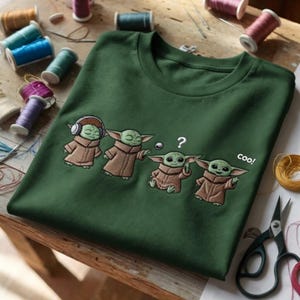 Puede incluir: Camiseta verde oscuro con un dise&ntilde;o bordado de cuatro personajes de Grogu en varias poses. Uno lleva auriculares, otro tiene un signo de interrogaci&oacute;n y el &uacute;ltimo dice "coo!"